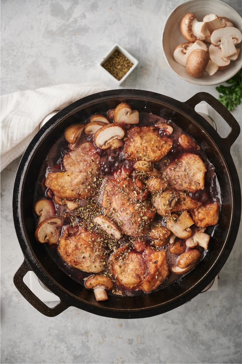 A cast iron pan with cooked chicken marsala in the pan and a small bowl of raw mushrooms and oregano placed next to the pan.