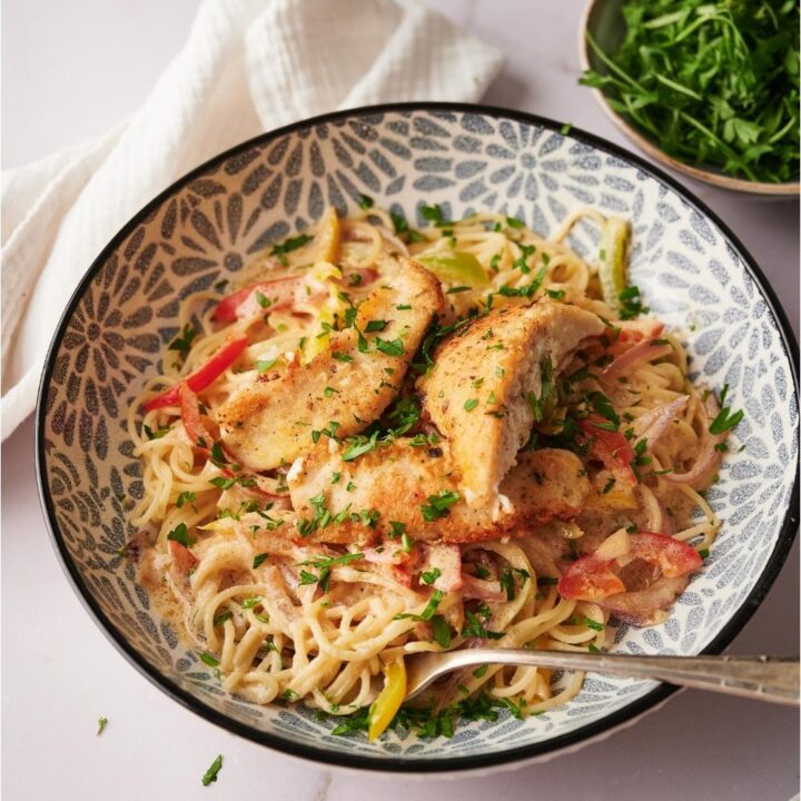 Chicken scampi on a blue and white decorated plate with a fork in the scampi and a bowl of parsley and a white dinner napkin next to the scampi, all on a grey counter.