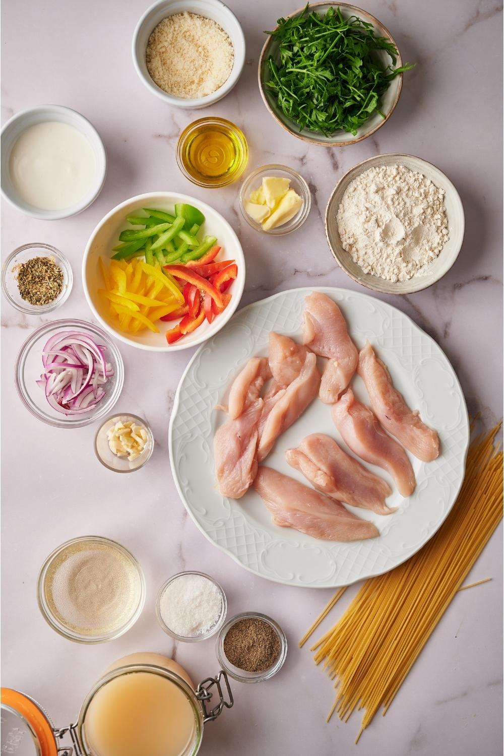 An assortment of ingredients including plates and bowls of raw chicken, flour and seasoning mixture, sliced bell peppers, sliced red onion, oil, butter, cheese, salt, pepper, parsley, and white wine, with raw pasta on the counter.