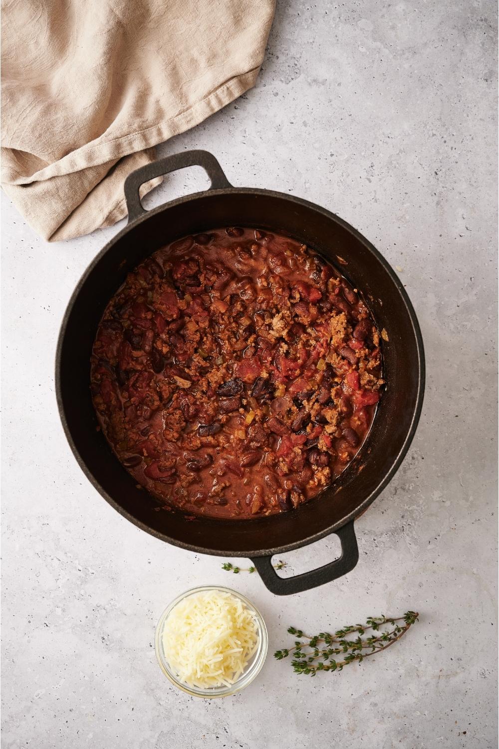 A large pot of chili on a grey counter that is surrounded by a white kitchen towel, a small bowl of cheese, and sprigs of dried thyme.