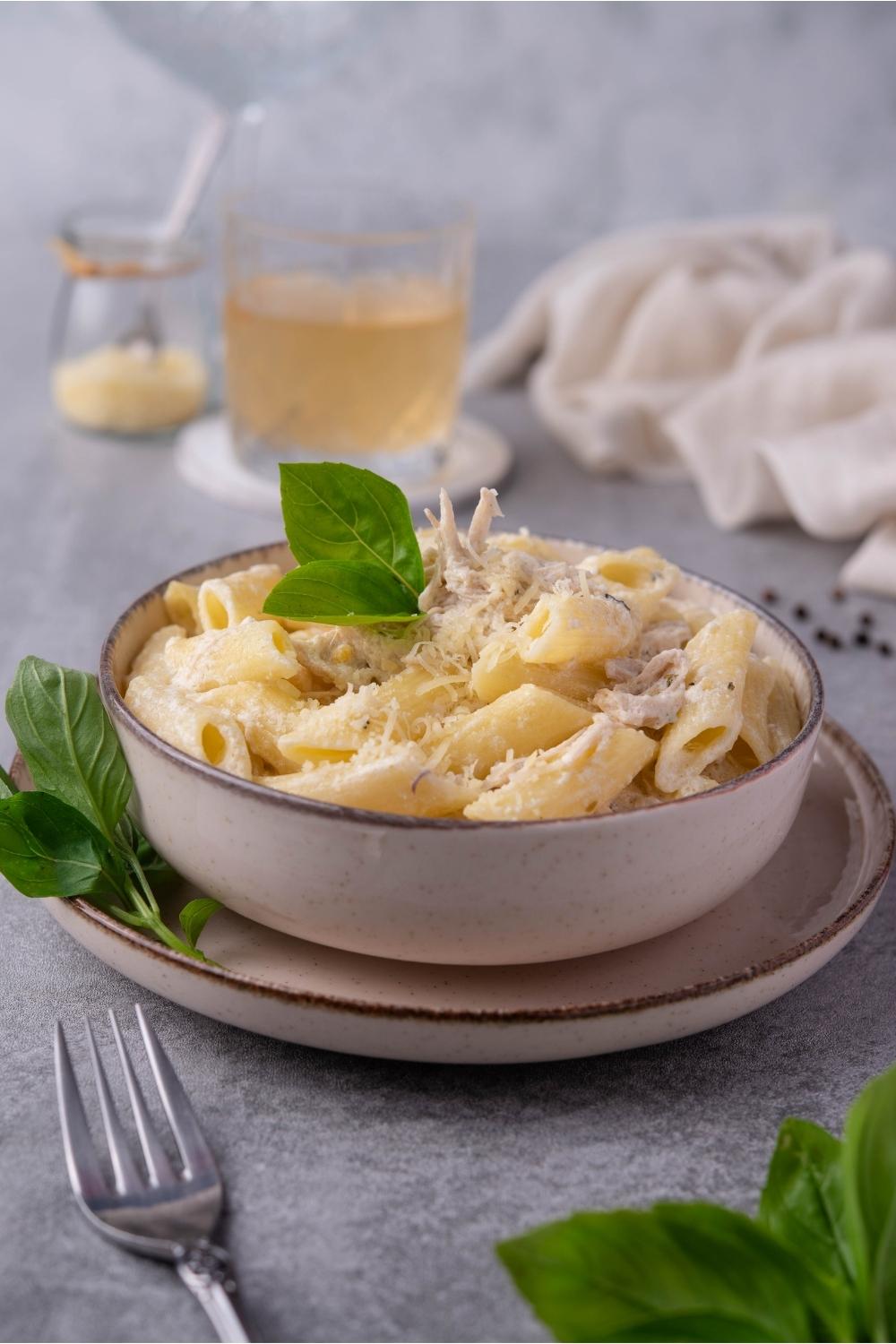 Olive Garden chicken pasta in a white bowl that is on a white plate. There is a fork next to the plate and basil leaves decorated around the pasta. In the background there is a full glass, a bowl of shredded cheese, and a white kitchen towel.