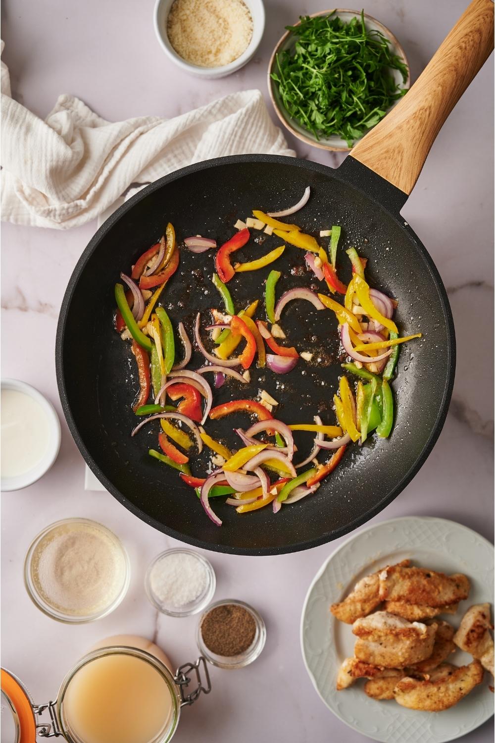 Skillet containing cooked sliced bell peppers, red onion, and garlic. Surrounding the pan are plates of ingredients including cooked chicken, salt, pepper, parsley, and cheese.
