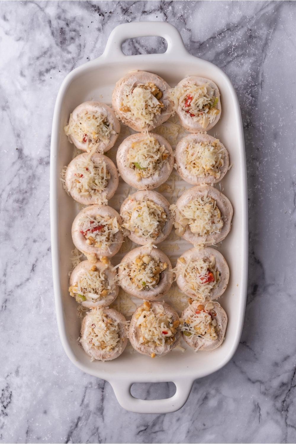 A white baking dish filled with uncooked stuffed mushrooms covered in cheese.