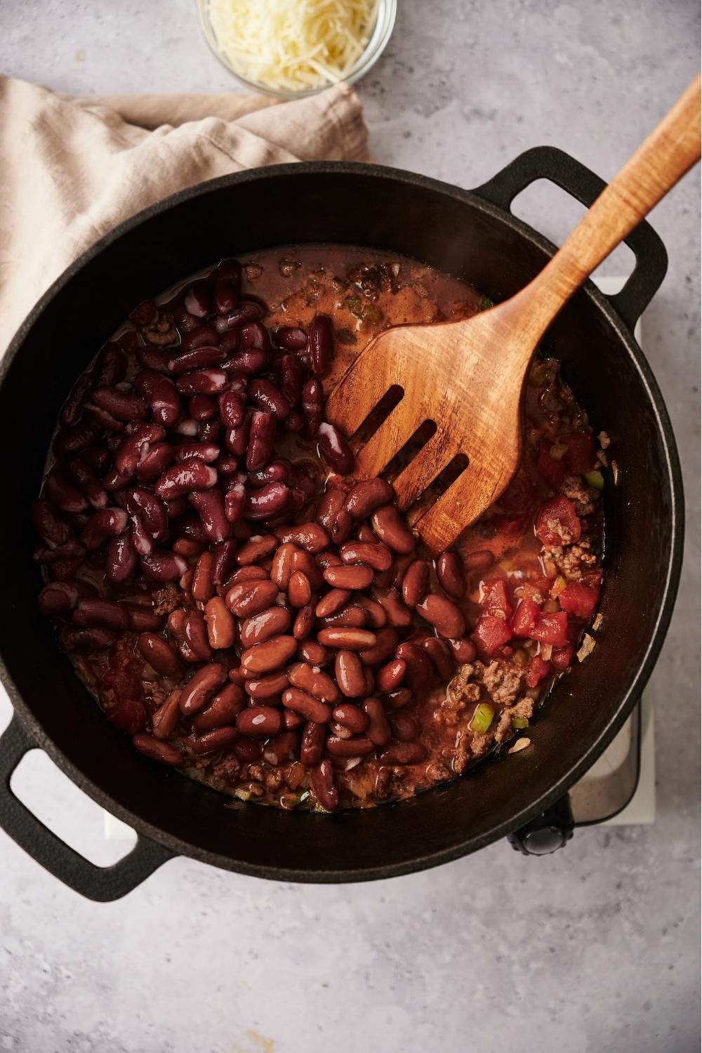 A large pot with chili simmering in the pot and a slotted wooden spatula in the pot. Kidney beans and pinto beans have just been added to the pot and have not yet been stirred. The pot sits on a grey counter next to a white kitchen towel and a small bowl of shredded cheese.