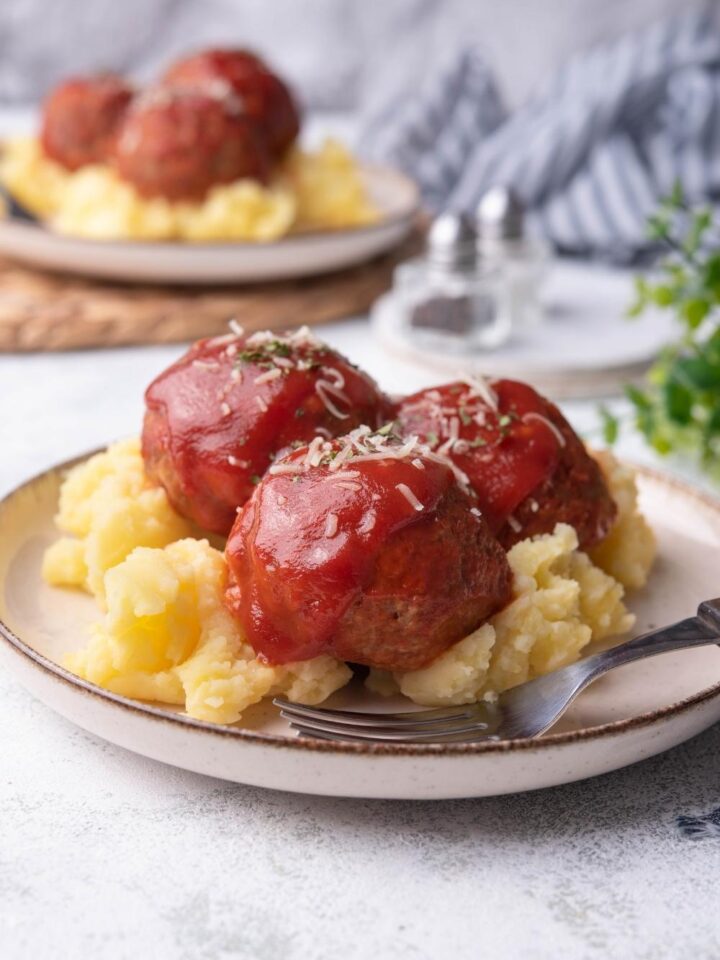 Ham balls on a white plate covered in tomato sauce on top of mashed potatoes with a fork on the plate, with a second plate of ham balls in the background.