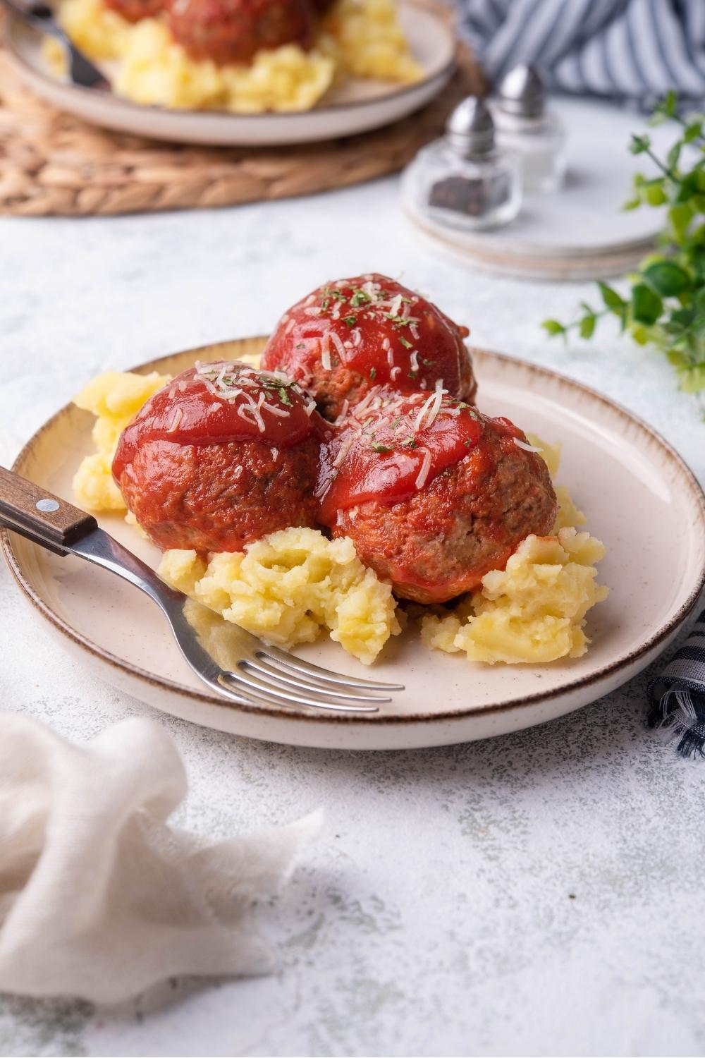 Ham balls on a white plate covered in tomato sauce on top of mashed potatoes with a fork on the plate, with a second plate of ham balls in the background.