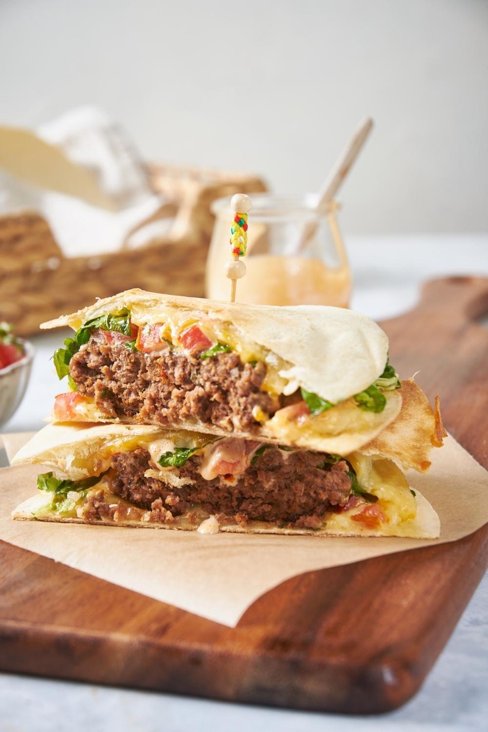 Quesadilla burger sliced in half, stacked on top of each other, on a piece of parchment paper on a wood cutting board, with a toothpick stuck through the burgers. There is an empty jar of spicy mayo behind the burgers.