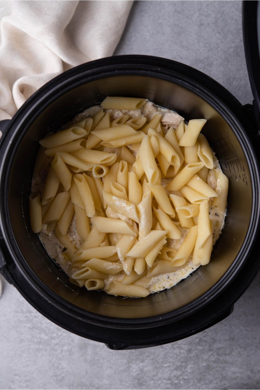 A black slow cooker with shredded chicken, creamy Italian dressing, and freshly added cooked pasta in the slow cooker. The slow cooker is on a grey counter with a partial view of a white kitchen towel.