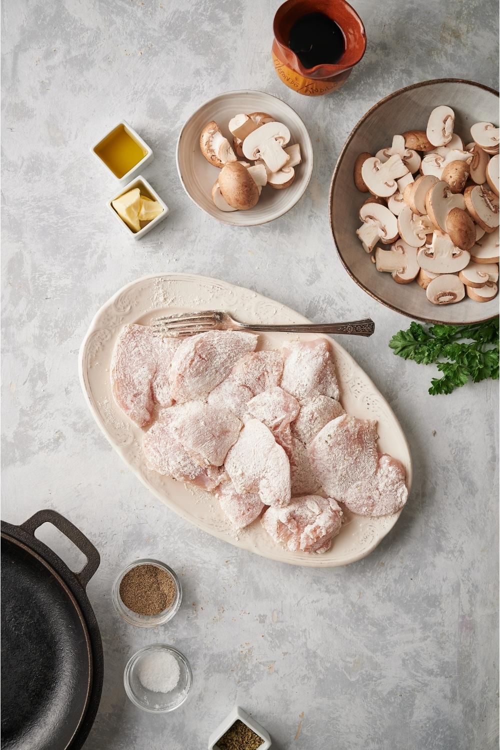 Raw chicken covered in a flour mixture on a white plate with a fork on the plate and small bowls of sliced mushrooms, salt, pepper, butter, oil, and marsala wine.