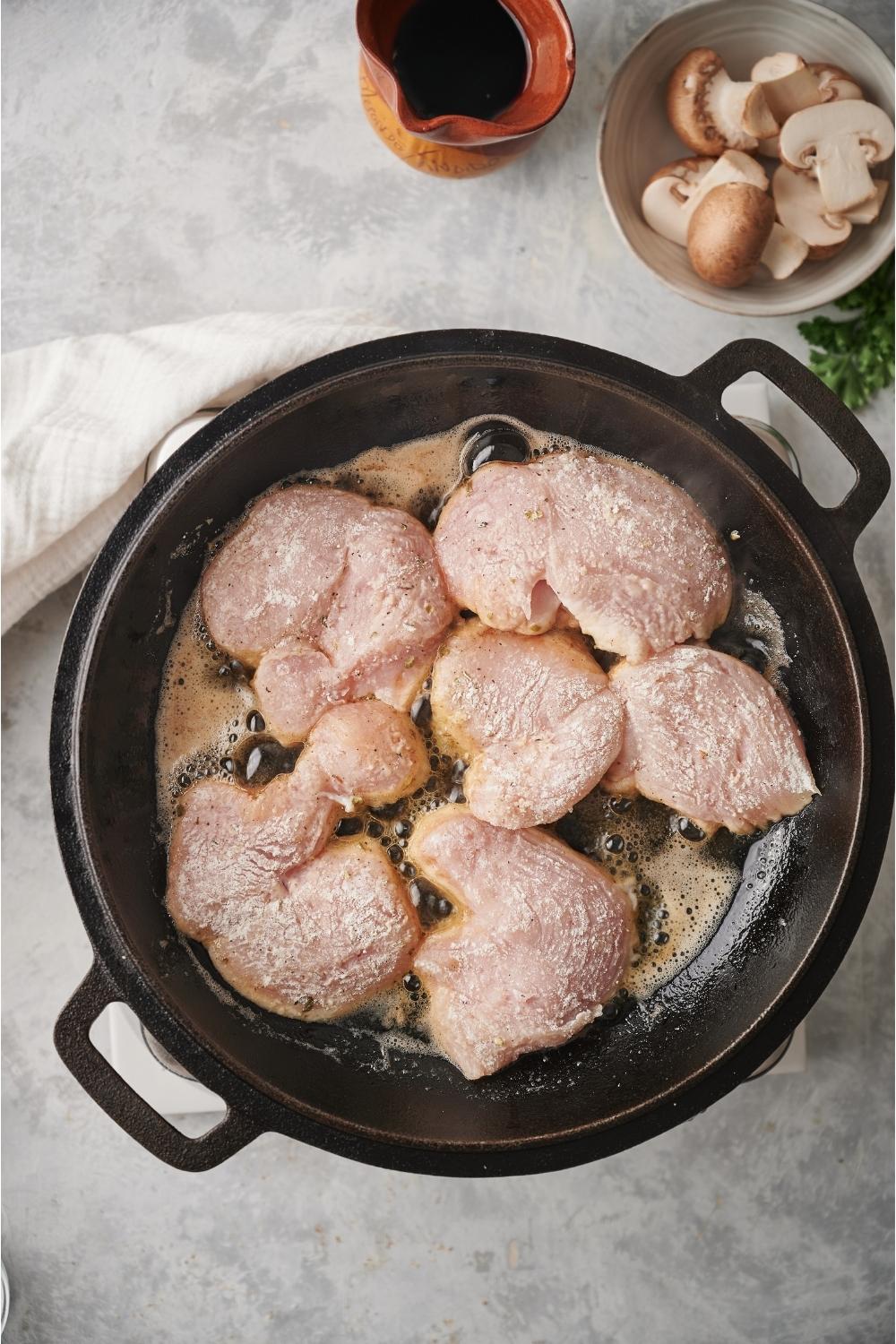 Raw chicken breast pieces covered in a flour mixture in a cast iron pan, with bubbling butter and oil and raw sliced mushrooms in a bowl next to the pan.