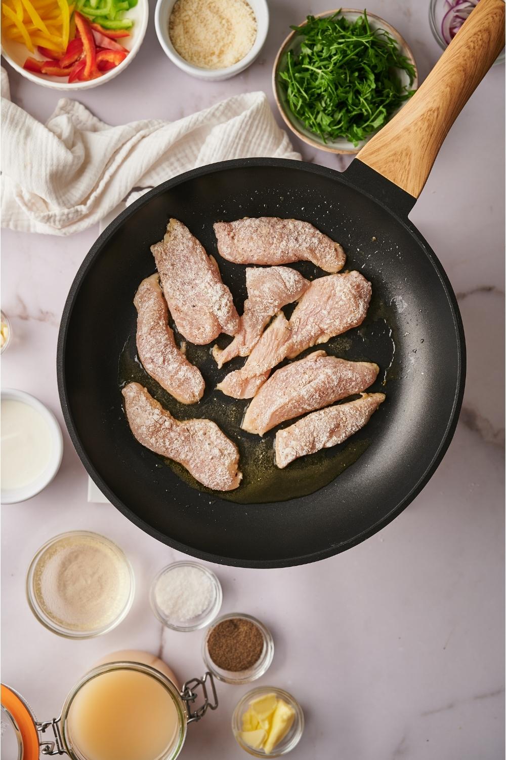 A skillet cooking raw chicken that has been tossed in a flour and seasoning mixture, with raw ingredients surrounding the skillet including bowls of sliced bell peppers, cheese, butter, oil, garlic, and parsley.