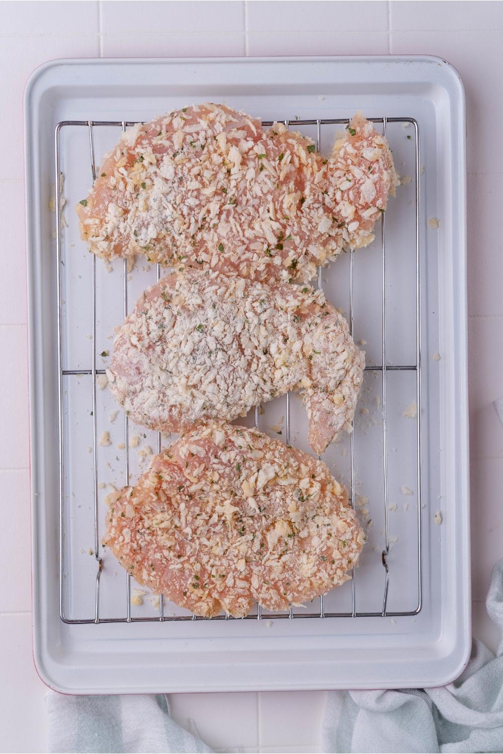 Raw flattened chicken breasts coated in a bread crumb mixture laid out on a rack that's on a baking sheet.