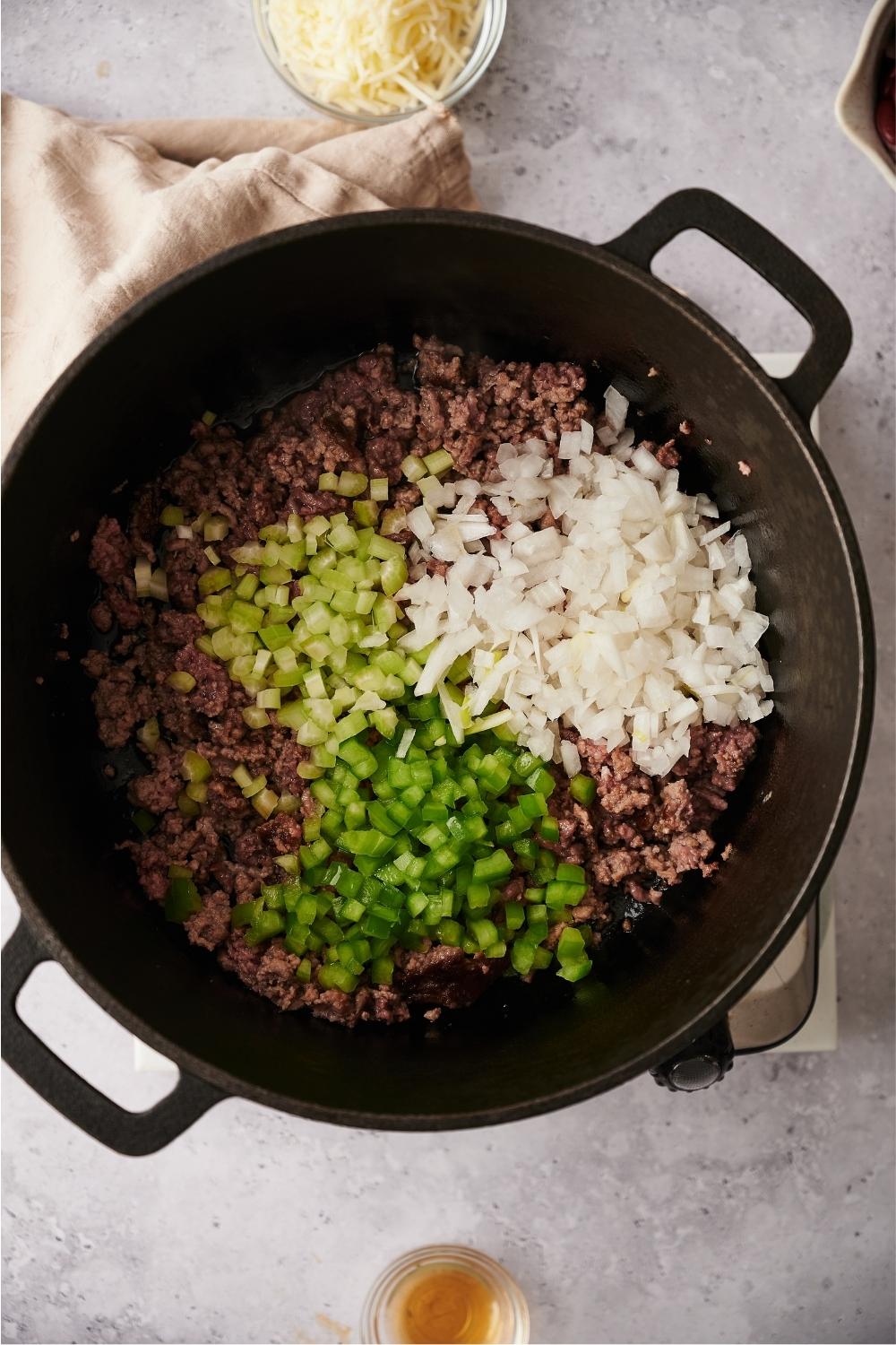A large pot containing ground beef, green bell pepper, onion, and celery. The pot sits on a grey counter and is surrounded by a kitchen towel and bowls of cheese and vinegar.
