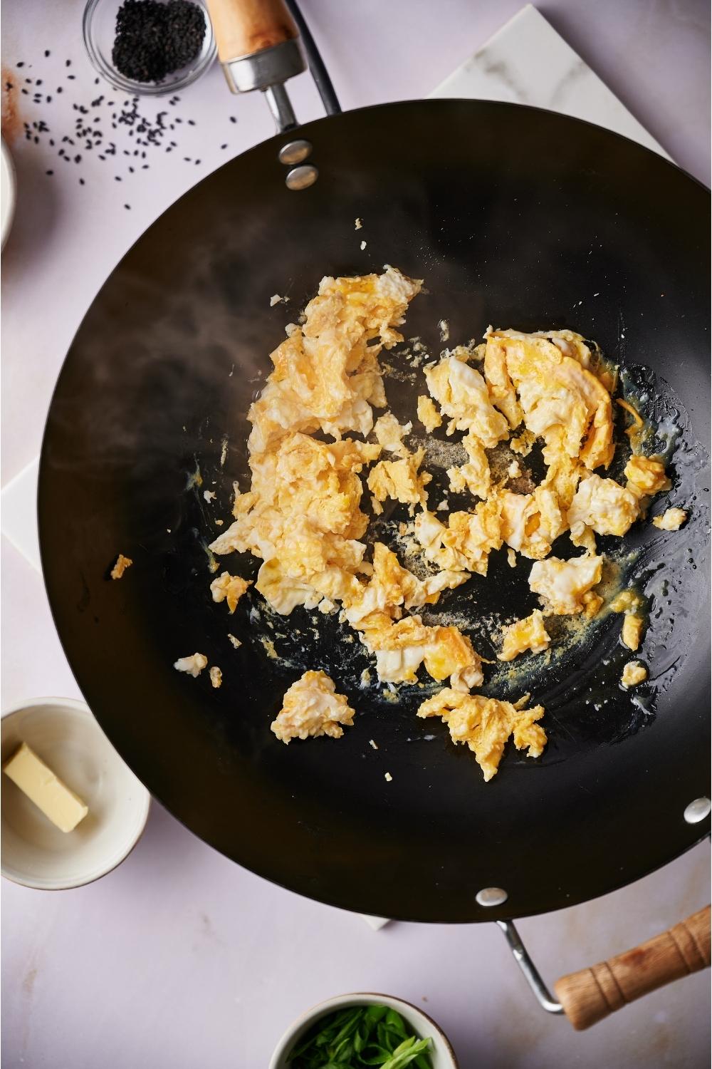 A wok with freshly scrambled eggs cooking. The wok is on a grey counter and there are bowls of sesame seeds, butter, and chopped green onions next to the wok.