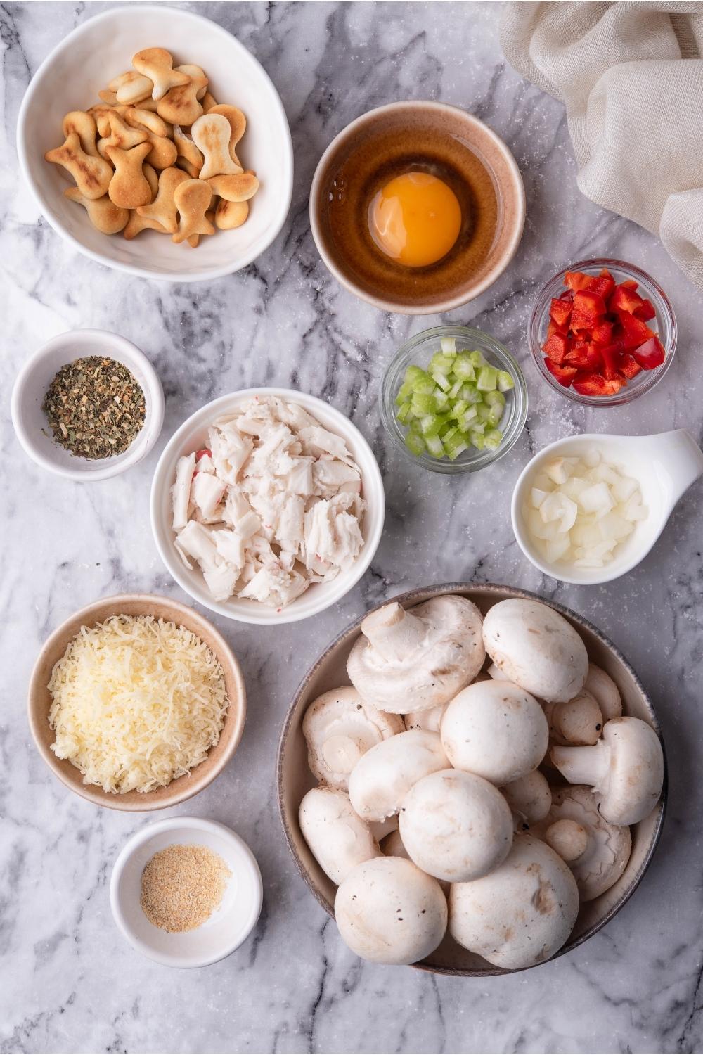 An assortment of ingredients for stuffed mushrooms including bowls of crackers, mushrooms, egg, cheese, vegetables, and spices.