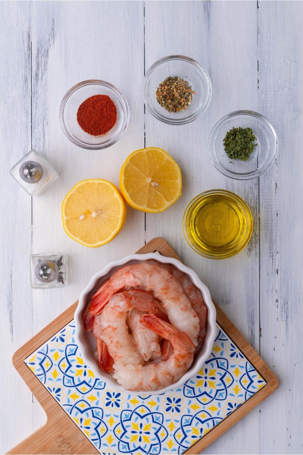 An assortment of ingredients for grilled shrimp including bowls of raw shrimp, seasonings, oil, and a lemon sliced in half.