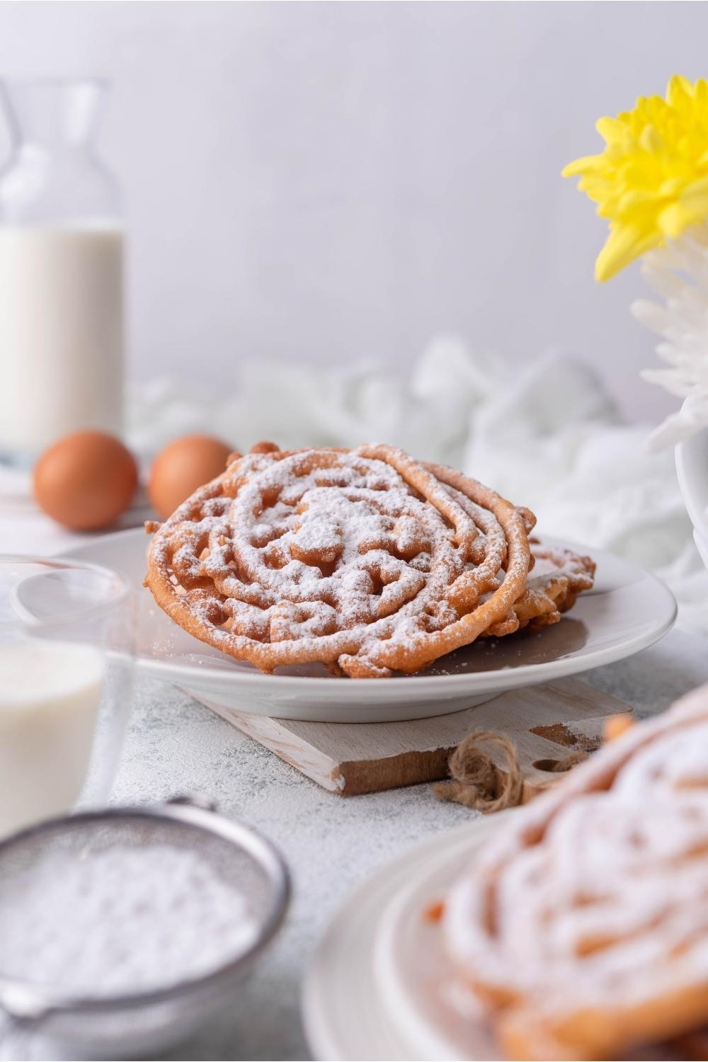 A plate of funnel cakes covered in powdered sugar, with ingredients surrounding the plate.