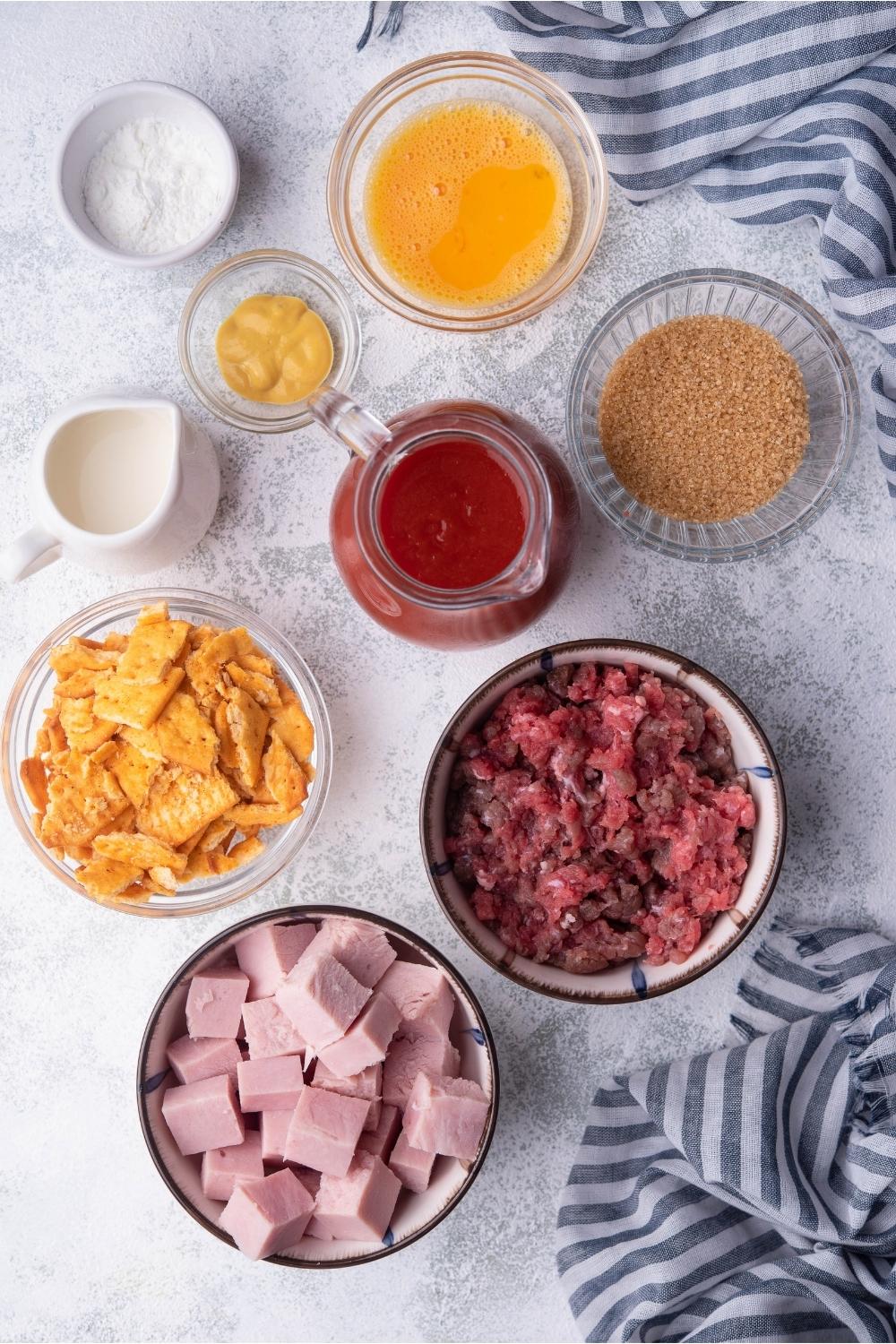 An assortment of ingredients for ham balls including bowls of tomato sauce, egg mixture, cubed ham, ground beef, saltines, and seasonings.