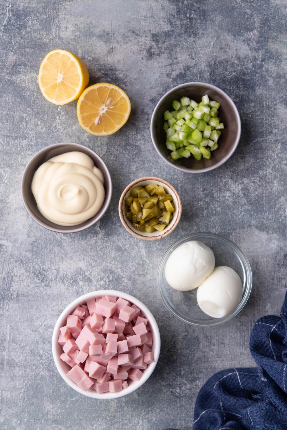 An assortment of ingredients for ham salad including bowls of diced ham, hard boiled eggs, lemon, mayonnaise, pickles, and celery.