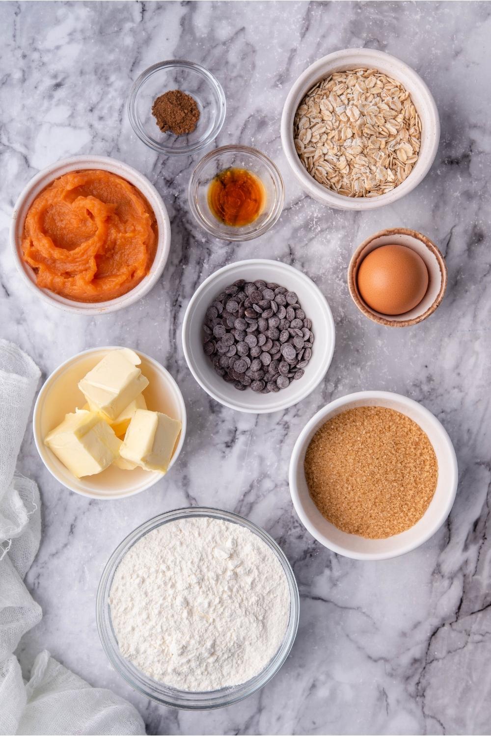 An assortment of ingredients for sweet potato cookies including bowls of sweet potato mash, butter, flour, chocolate chips, eggs, brown sugar, vanilla extract, and spices.