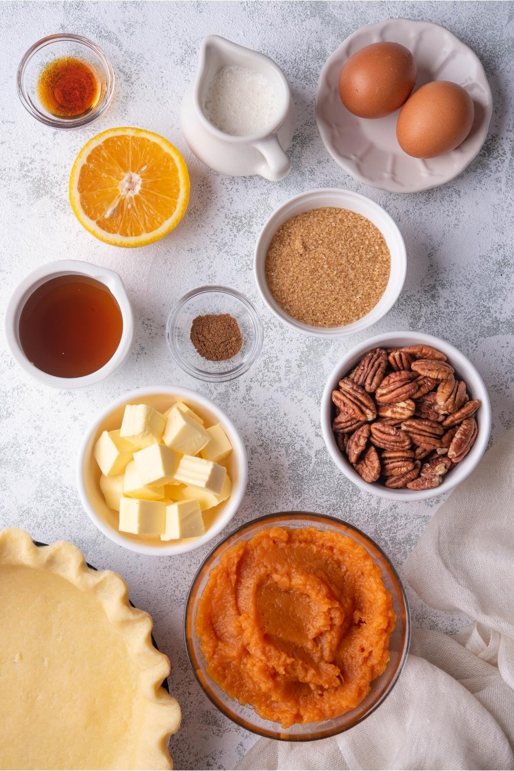 An assortment of ingredients for sweet potato pecan pie including bowls of butter, pecans, mashed sweet potato, sugar, maple syrup, and an unbaked pie crust.