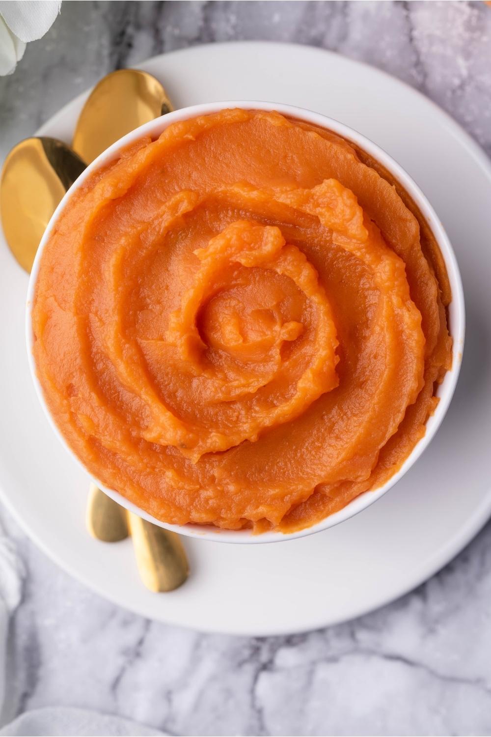 Sweet potato puree in a white bowl on a white plate with two spoons.