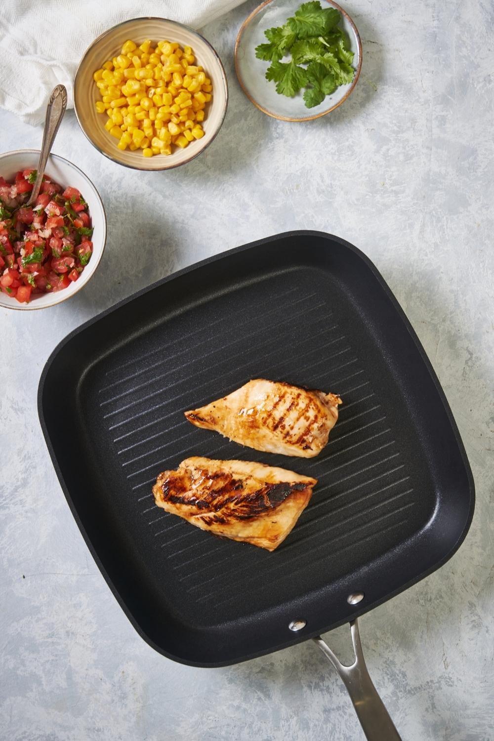A grill pan with grilled chicken breasts on a grey counter surrounded by bowls of corn, cilantro, and pico.
