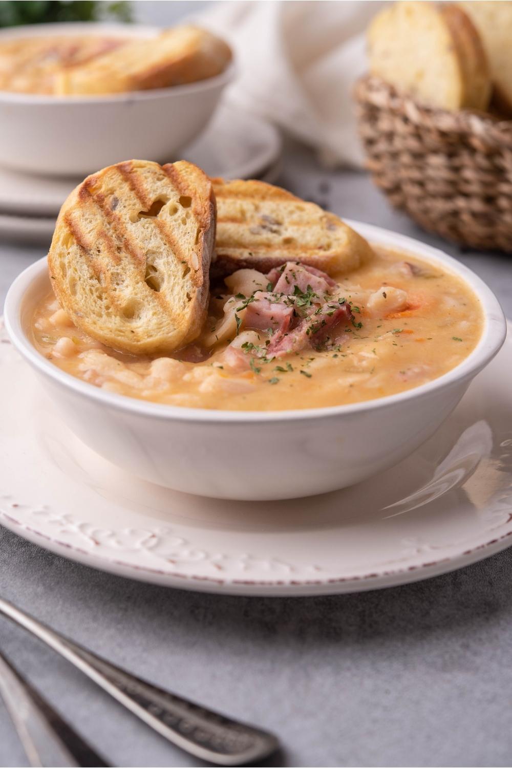 Ham and beans soup in a white bowl with toasted bread on top and a spoon next to the bowl. A second bowl of soup is in the background.