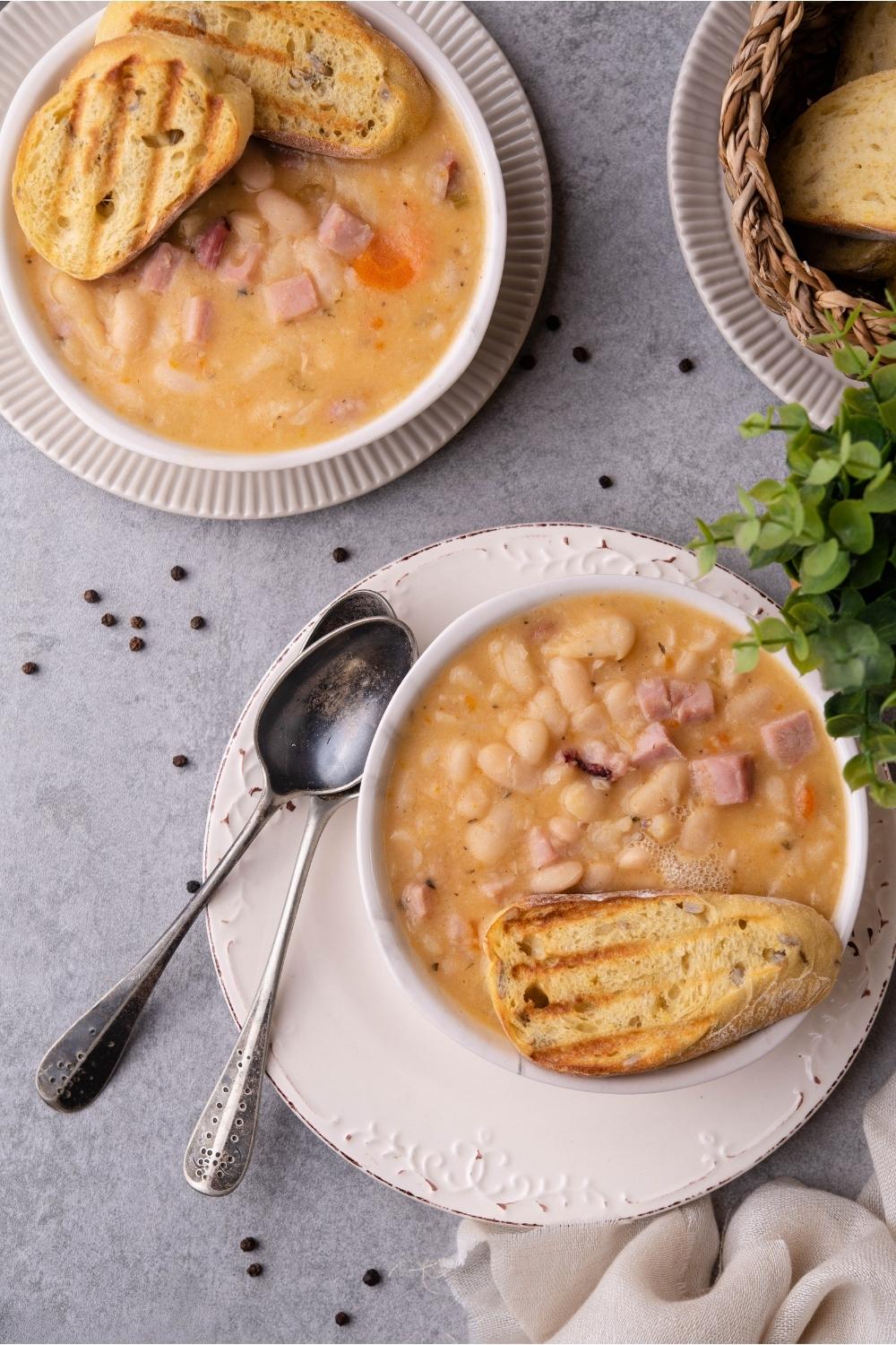 Two bowls of ham and beans soup, each topped with toasted bread. Two spoons lay next to one of the bowls of soup.