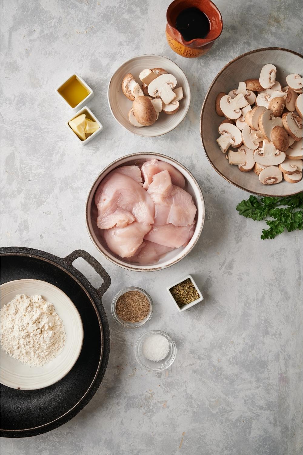 A variety of ingredients for chicken marsala including bowls of raw chicken, sliced mushrooms, salt, pepper, butter, and a flour mixture, all on a grey countertop.