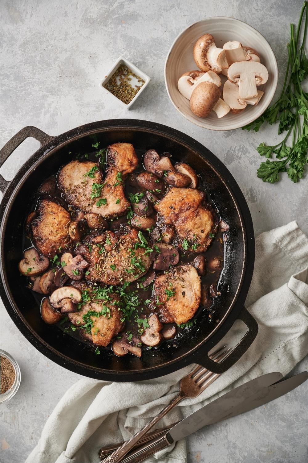 Cooked chicken marsala in a cast iron pan with a napkin, fork, knife, parsley sprigs, a bowl of raw mushrooms, and oregano all surrounding the pan.