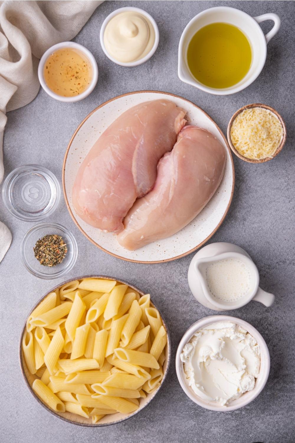 An assortment of ingredients for Olive Garden chicken pasta including raw chicken breasts, cooked pasta, cream cheese, shredded cheese, and mayo. Everything is on a grey counter and there is a partial view of a white kitchen towel.