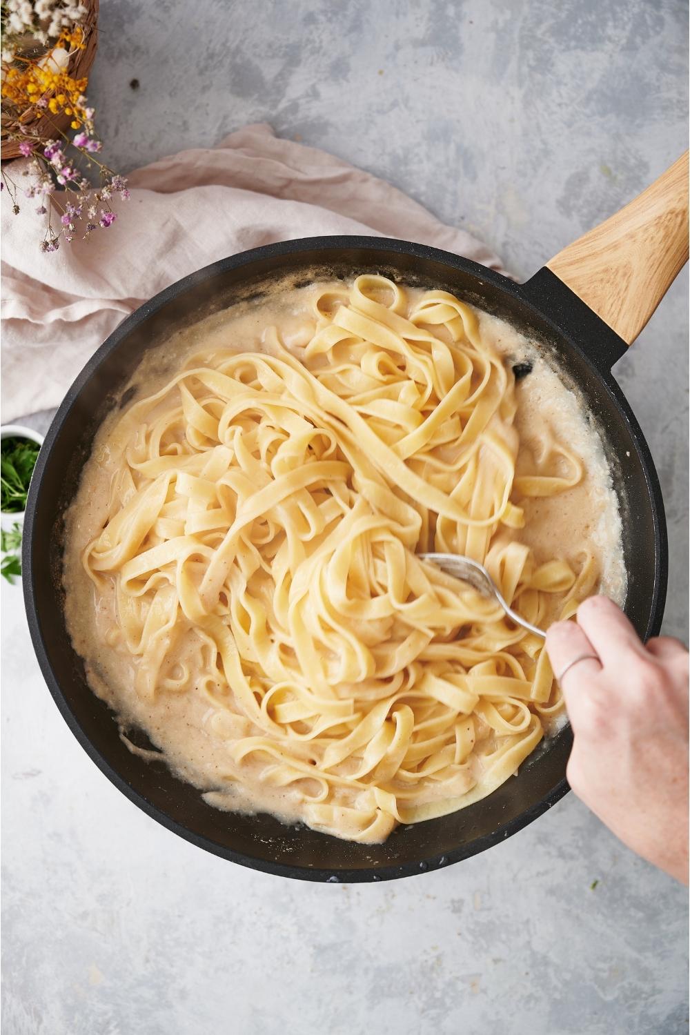 A pan with finished alfredo sauce and cooked noodles being stirred up with a hand holding a spoon.