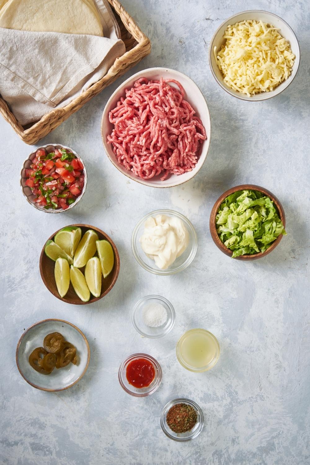 An assortment of ingredients including bowls of raw beef, pico, cheese, lettuce, limes, mayo, chili sauce, and a basket of tortillas.