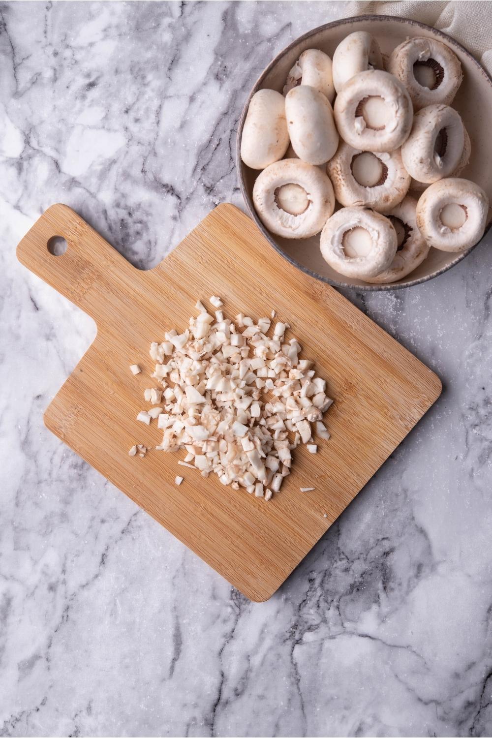 Chopped mushroom stems on a wood cutting board with a bowl of stemless mushrooms next to the board.