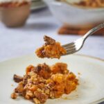 A fork picking up a piece of Ruth's Chris sweet potato casserole, and a baking dish of casserole in the background.