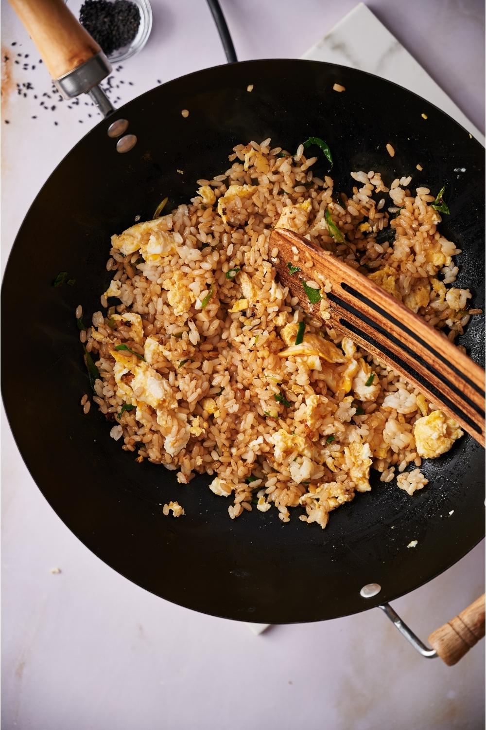 A wok with hibachi fried rice cooking and a spatula mixing the rice together. The wok is on a grey counter. Next to the wok there is a bowl of sesame seeds with seeds spilling out of the bowl.
