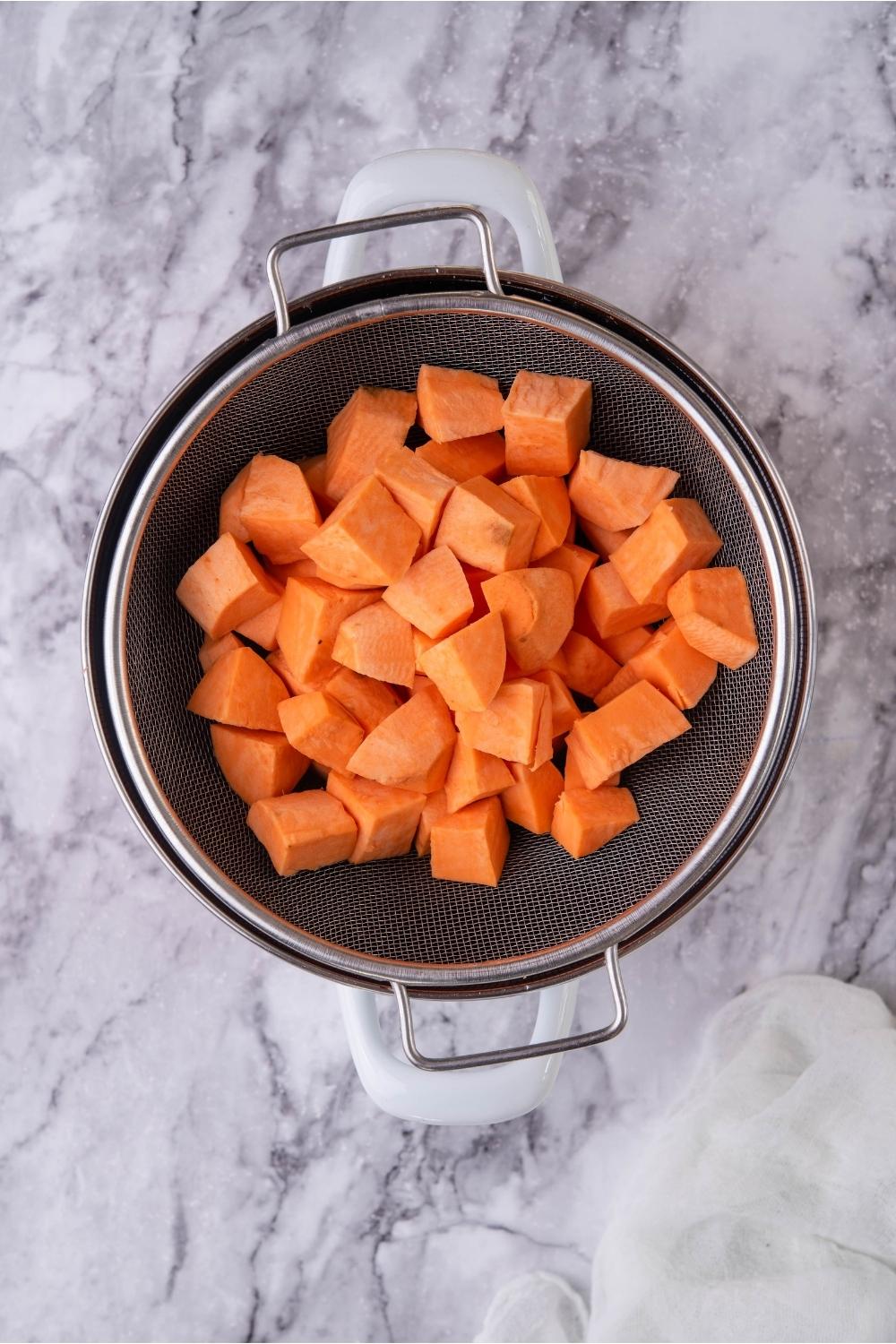 Uncooked cubes of sweet potato in a strainer that's over a large pot.