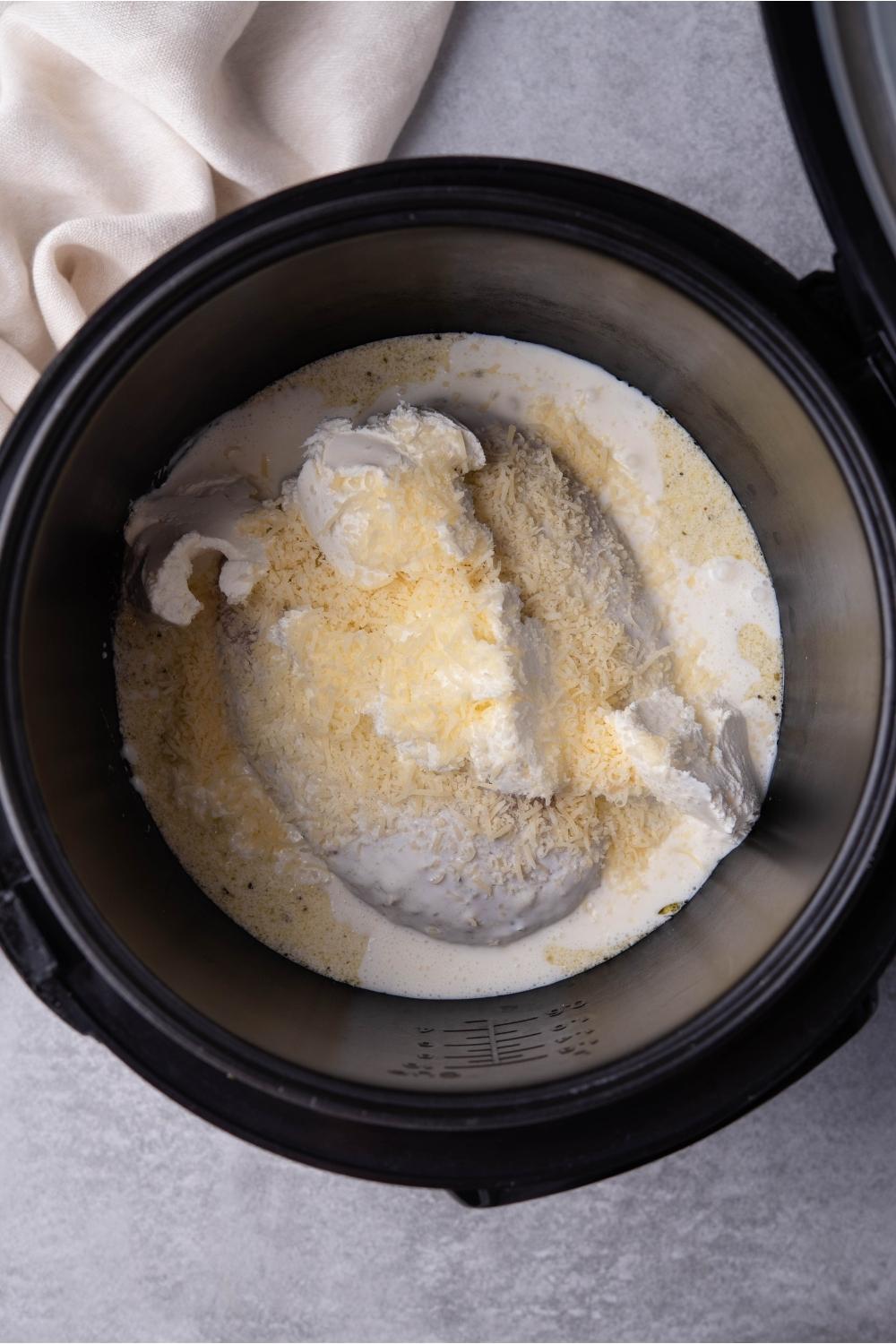 A black slow cooker with cooked chicken breasts covered in a creamy Italian dressing and shredded cheese. The slow cooker is on a grey counter and there is a partial view of a white kitchen towel.