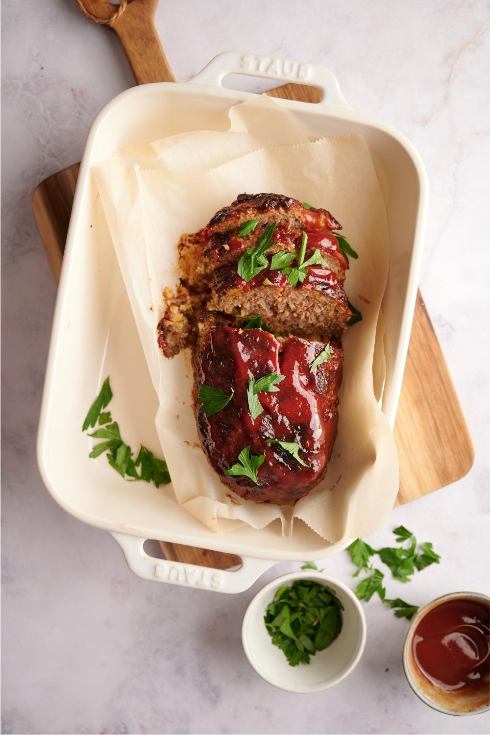 Southern meatloaf on parchment paper in a white baking dish. The dish is resting on a wood cutting board. Half of the meatloaf has been sliced to serve and the meatloaf is garnished with fresh parsley. Half-empty bowls of parsley and ketchup lay next to the meatloaf.