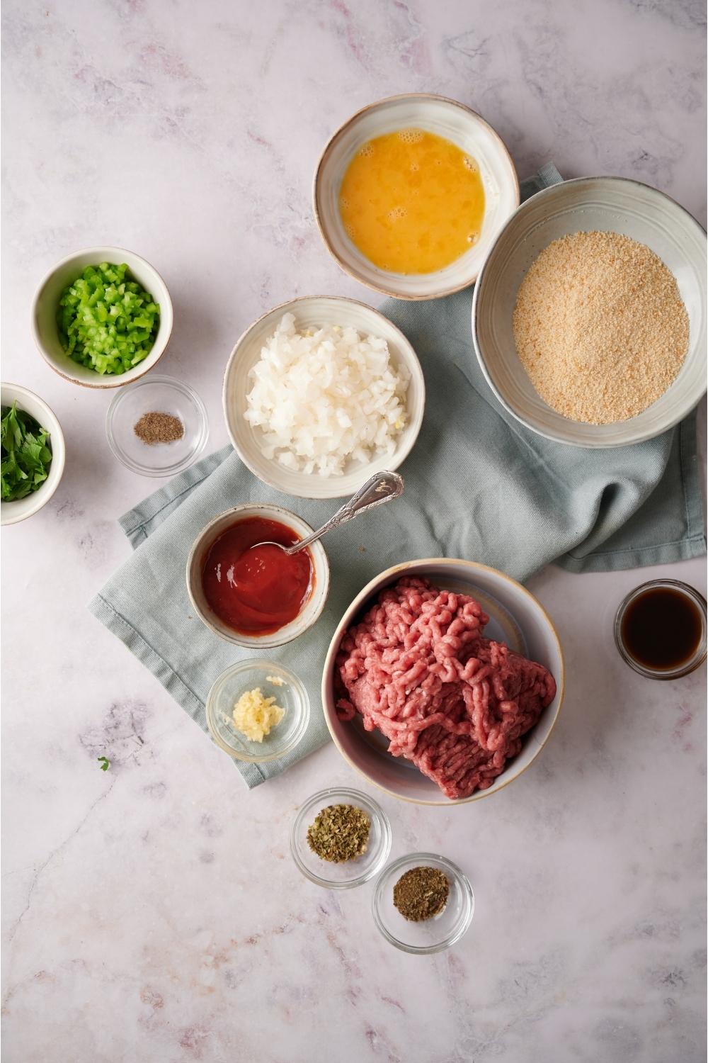 An assortment of ingredients including bowls of bread crumbs, egg, chopped onion, raw ground beef, ketchup, and seasonings. Most of the bowls are on a blue dish towel and everything is on a grey counter.