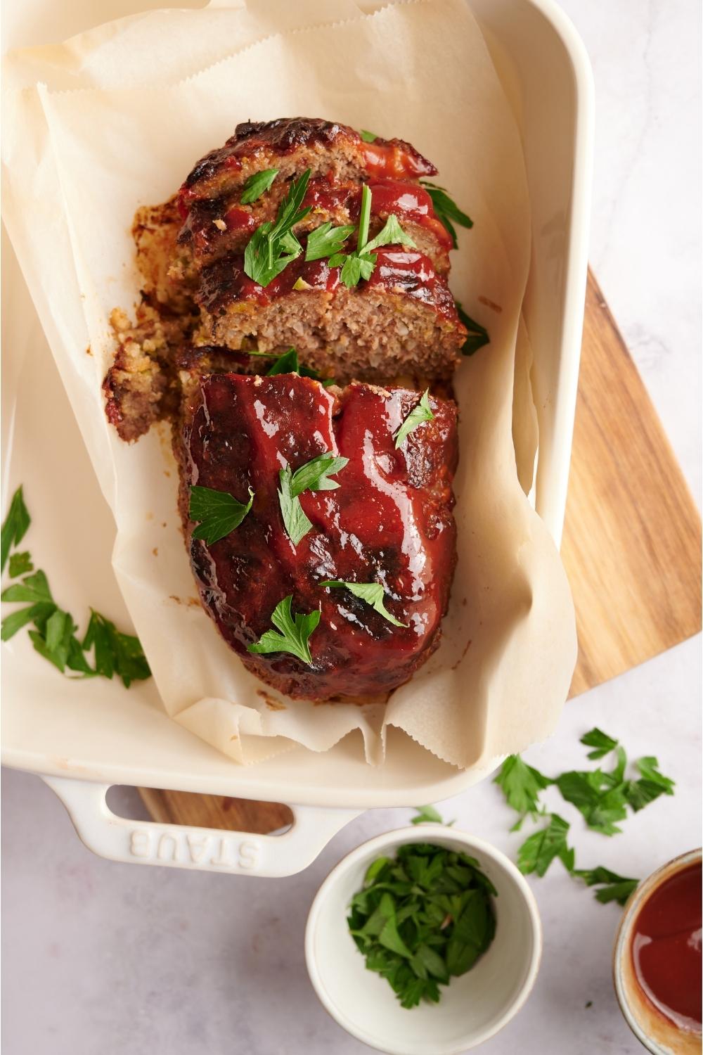 Southern meatloaf on parchment paper in a white baking dish. The dish is resting on a wood cutting board. Half of the meatloaf has been sliced to serve and the meatloaf is garnished with fresh parsley. A half-empty bowl of parsley is next to the meatloaf.