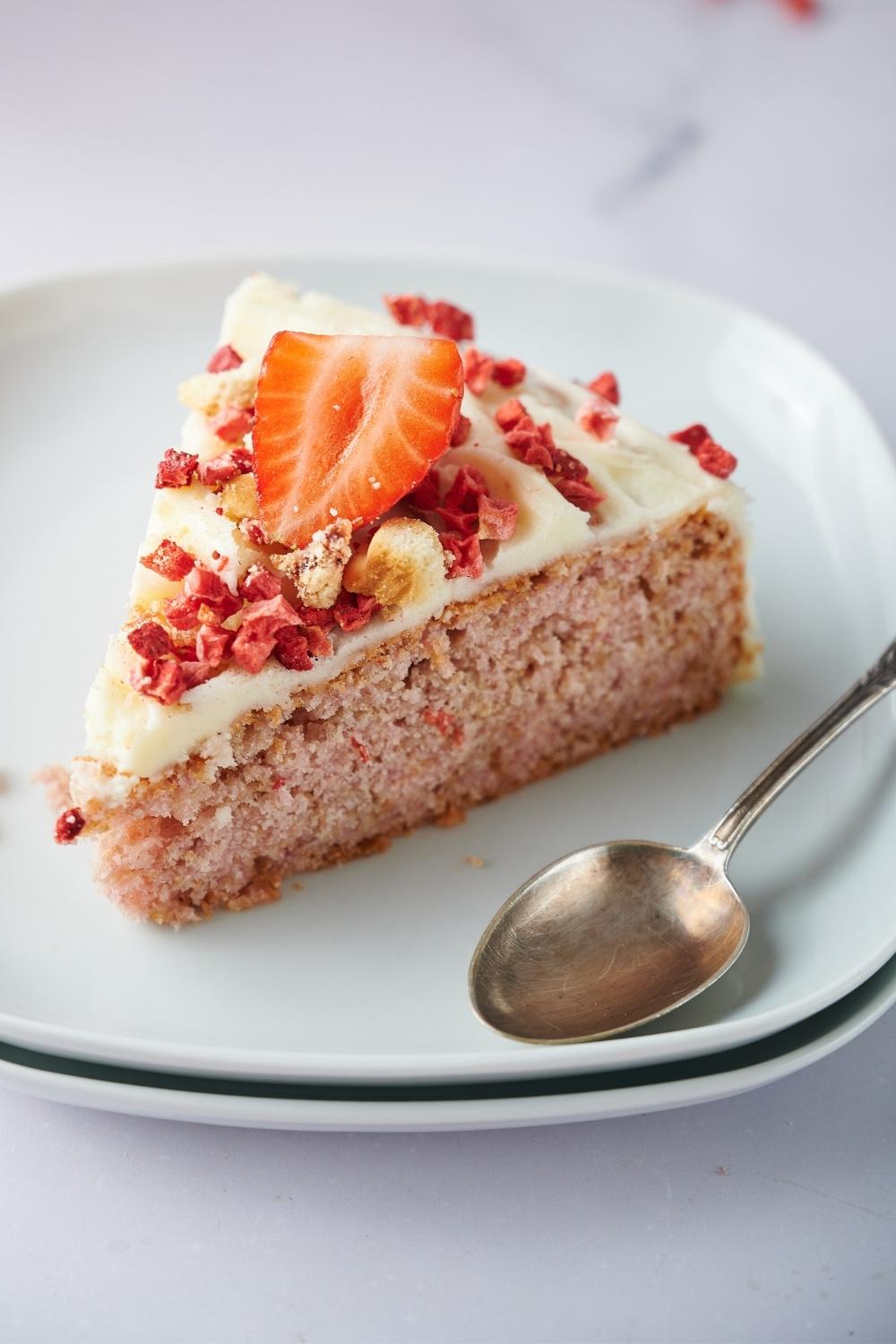 A slice of strawberry crunch cake toped with dried strawberry crumbles, frosting and a fresh strawberry slice on a plate. A spoon lays across the plate.