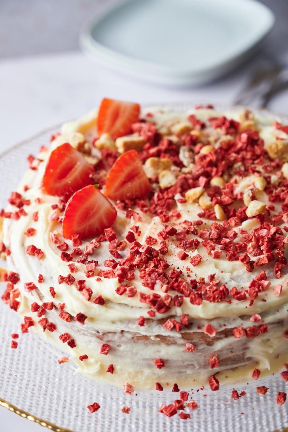 A close up of a whole strawberry cake on a serving plate. It's topped with dried strawberries, crumbled cookies, frosting and fresh sliced strawberries.