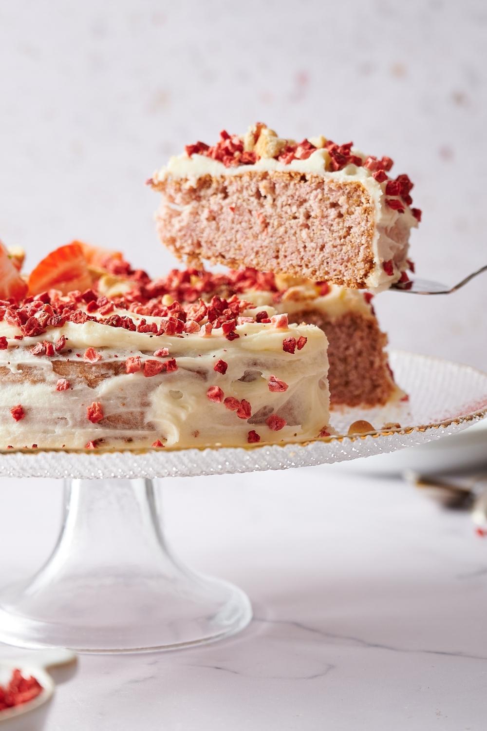 On a cake stand sits a whole strawberry cake on a serving plate. It's topped with dried strawberries, crumbled cookies, frosting and fresh sliced strawberries. A slice has just been cut and a spatula has lifted it above the cake.