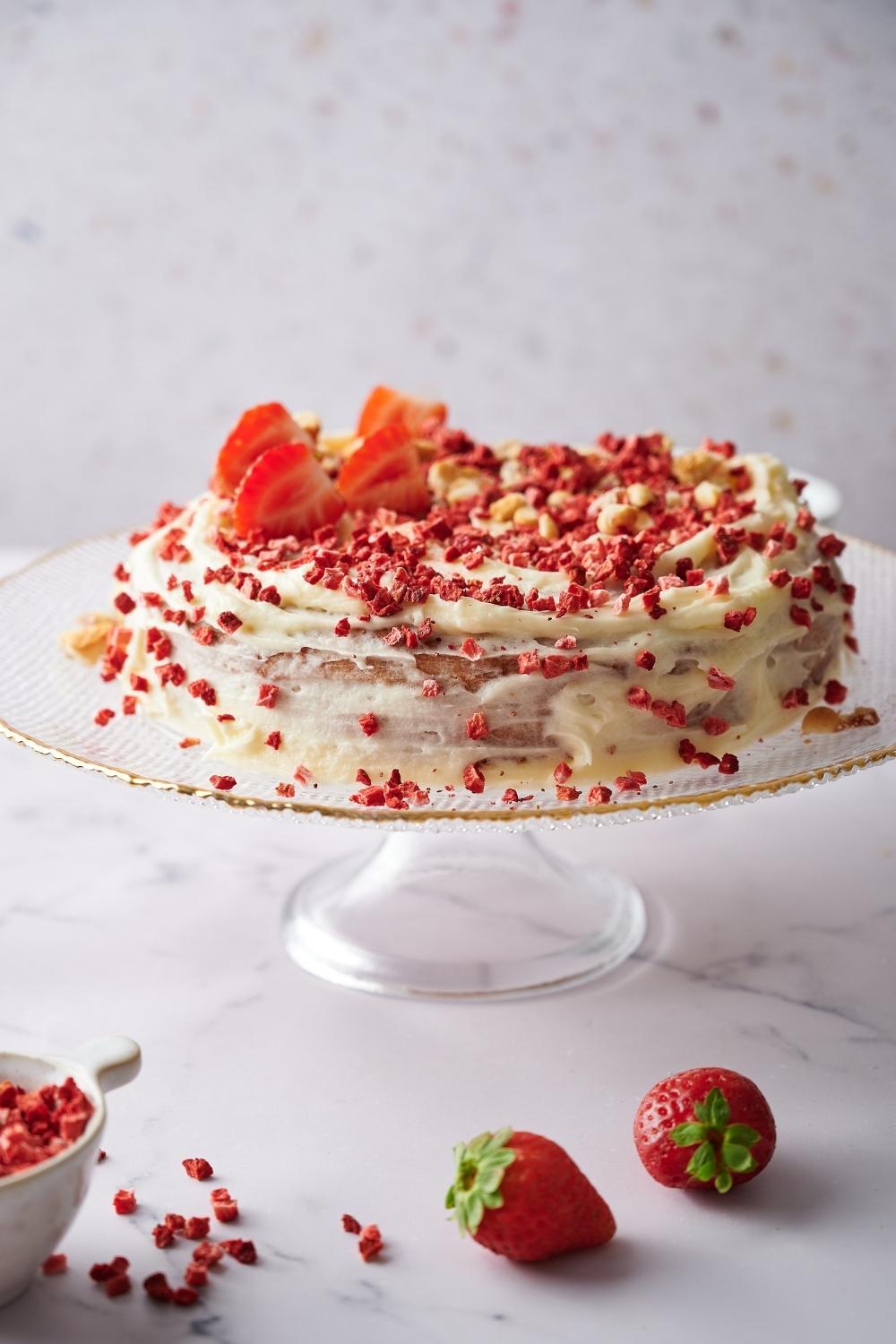On a cake stand sits a whole strawberry cake on a serving plate. It's topped with dried strawberries, crumbled cookies, frosting and fresh sliced strawberries.