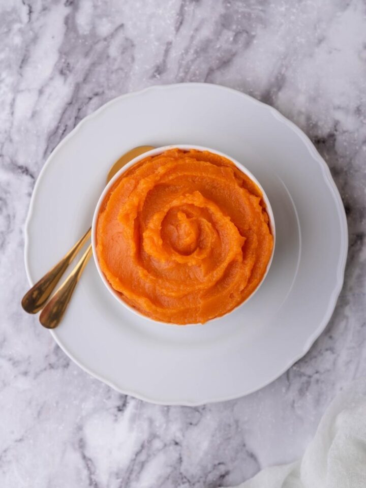 White bowl of sweet potato puree on a white plate with two spoons.