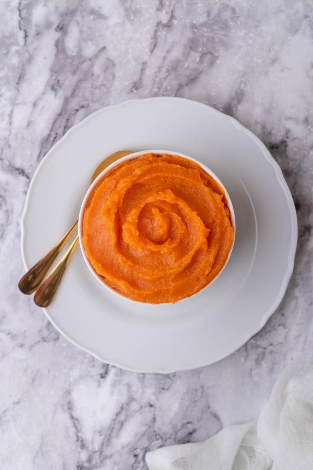 White bowl of sweet potato puree on a white plate with two spoons.