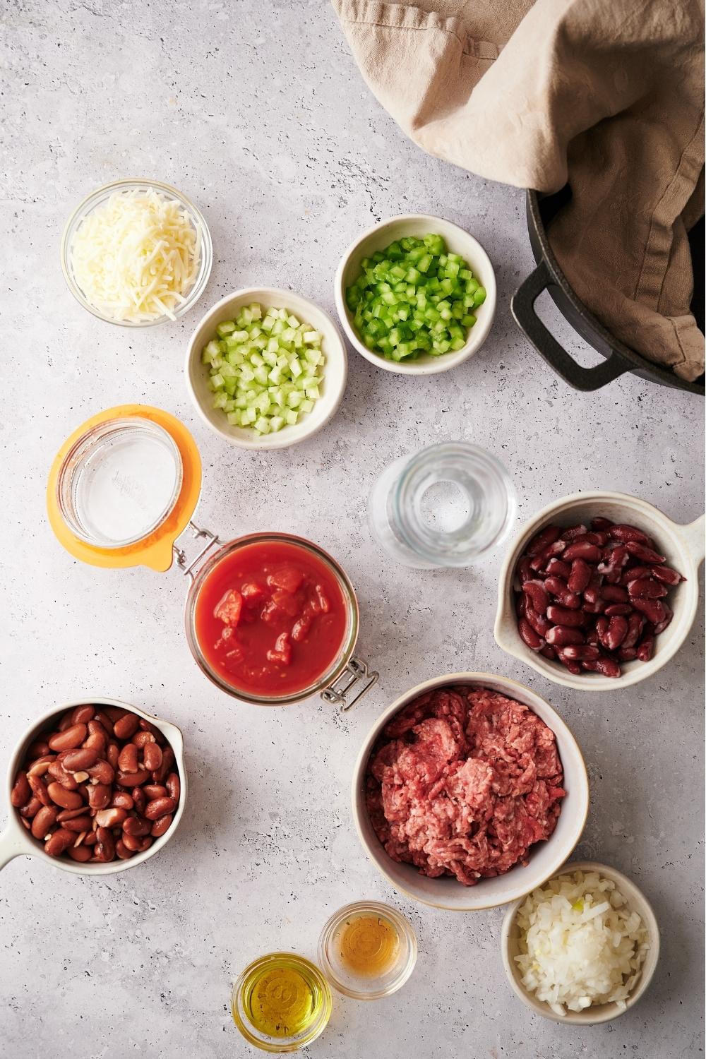 An assortment of ingredients for chili including bowls of stewed tomato, beans, raw ground beef, celery, onion, bell pepper, and more. There is a partial view of a large pot with a white kitchen towel in the pot.