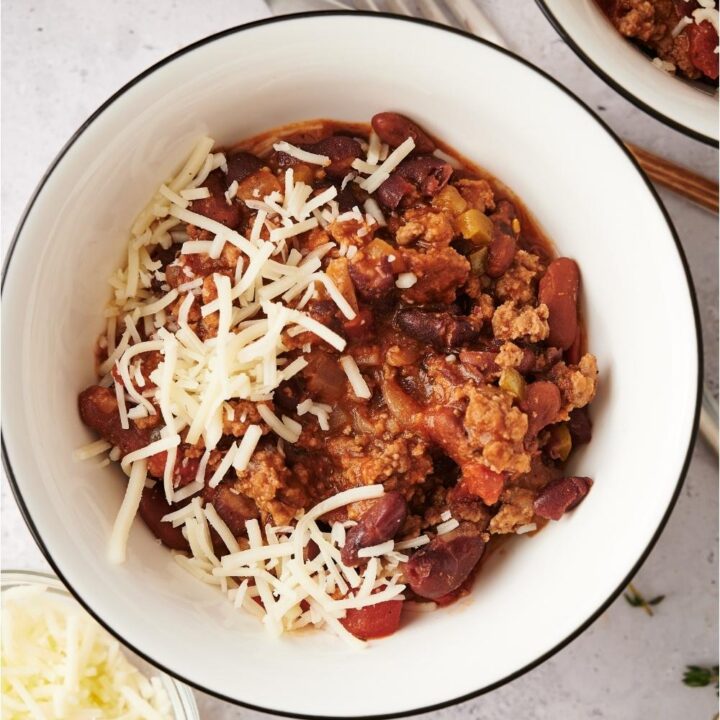 Wendy's chili in a white bowl on a grey counter, with a fork and knife in the background and partial views of a second bowl of chili, a small bowl of cheese, and a wooden board.